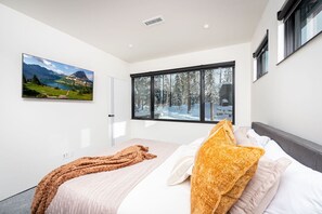 A modern bedroom with a large window, a bed with a white and orange blanket, and a painting of a mountainous landscape.