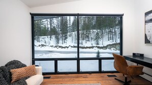 Ein modernes Zimmer mit großem Fenster und Blick auf einen verschneiten Wald, Holzboden, Sofa, Sessel und Schreibtisch mit Computer.