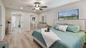 Une chambre à coucher comprenant un lit, un ventilateur de plafond, une fenêtre avec des stores et un couloir menant à une salle de bain.