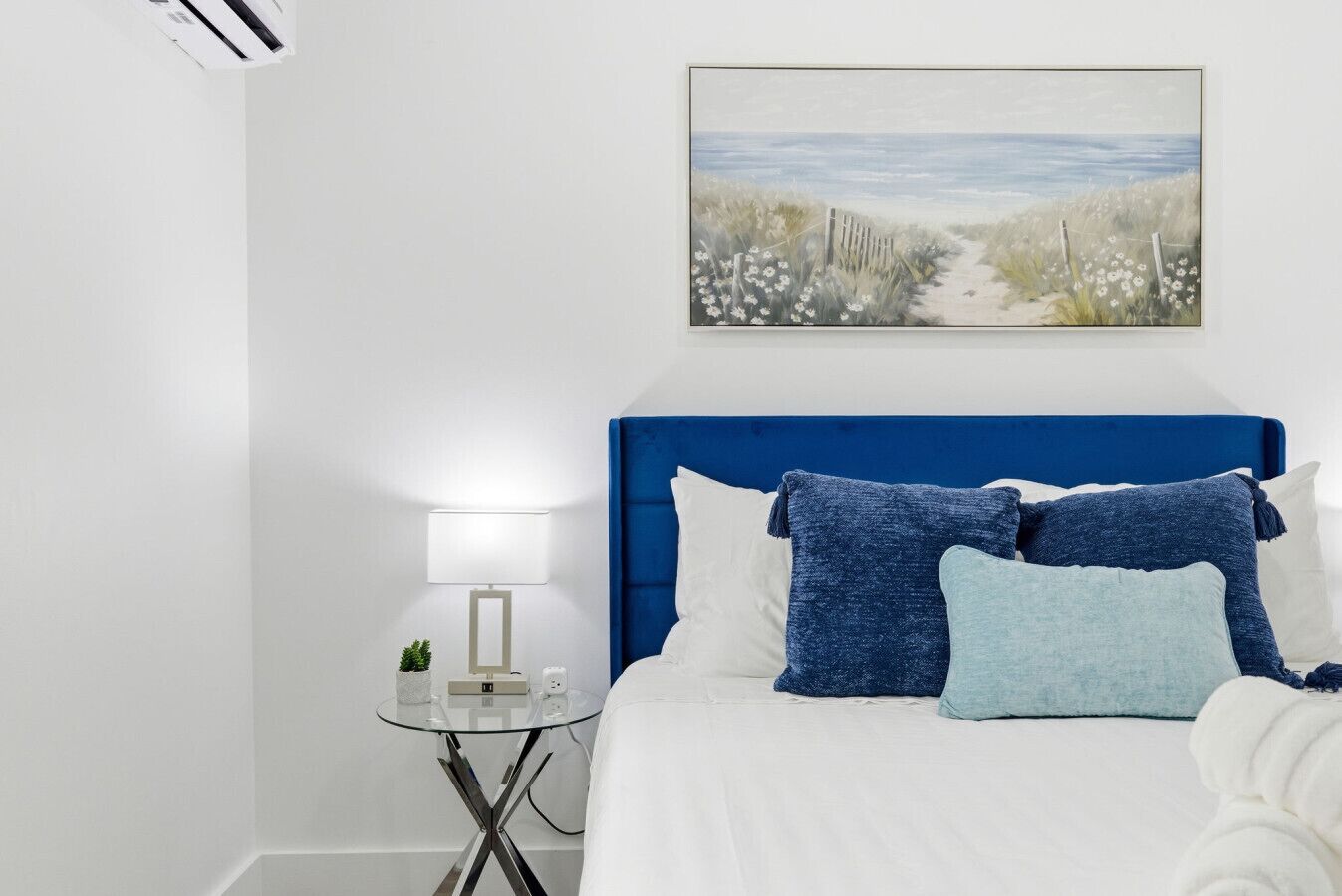 A bedroom with a white bed, blue headboard, and a painting of a beach scene.