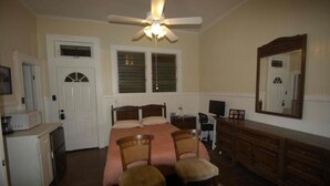 Ein Schlafzimmer mit einem Bett, zwei Stühlen, einem Schminktisch, einem Schreibtisch, einem Deckenventilator und einem Fenster mit Jalousien.