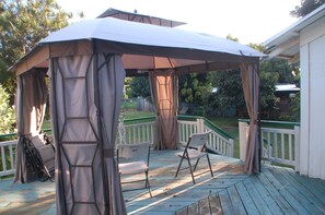 Een overdekt buitengebied met een houten terras, twee stoelen en een pergola.