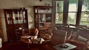 A spacious living room with a large wooden cabinet, multiple armchairs, a coffee table, and a view through large windows. - Hale Naalehu LLC Bed & Breakfast (Naalehu)