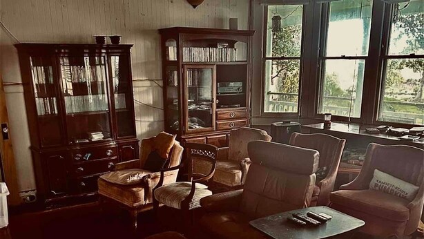 Ein geräumiges Wohnzimmer mit einer großen Holzkassette, mehreren Sesseln, einem Couchtisch und Blick durch große Fenster.