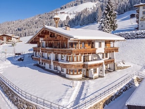 A large, multi-story chalet with a snow-covered roof and wooden balconies, surrounded by a snowy landscape and mountains.