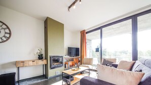 A modern living room with a fireplace, a television, and a large window with curtains. - Beautiful apartment overlooking the golf course (Barvaux)