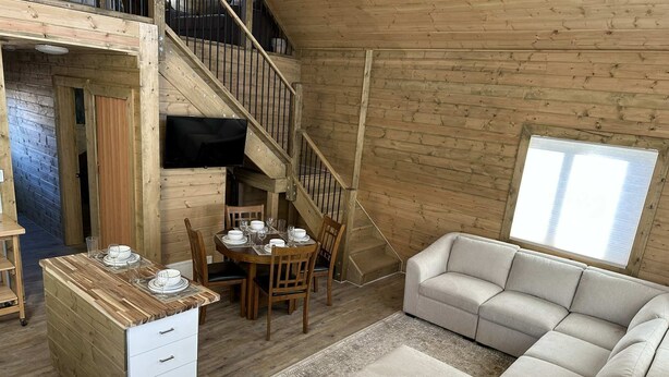A cozy loft-style room with a wooden staircase, a dining area with a table set for four, a sofa, and a TV mounted on the wall.