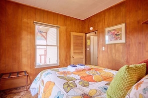 Ein Zimmer mit Holzwänden, einem Bett mit blumigem Überwurf, einem Fenster, einer Tür und einem gerahmten Bild an der Wand.