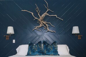 A bed with white linens and blue floral pillows, a wall-mounted tree branch decor, and two wall sconces with white lamps. - Sunflower Cottage Downtown (Pensacola)