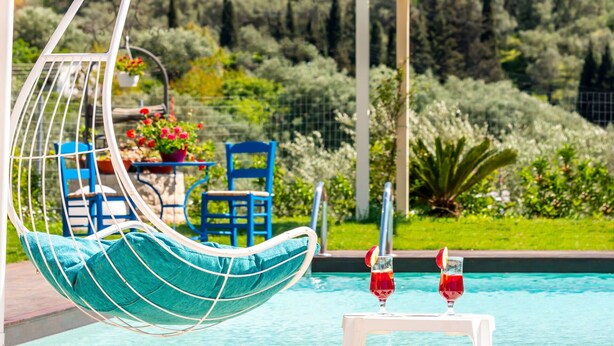 A poolside area with a hanging chair, two chairs, a table with drinks, and a view of a lush garden. - Villa Irida - Holiday home with pool 400 meters from Agios Spyridon beach in Corfu (Acharavi)