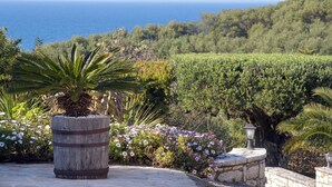 A landscaped garden with a wooden barrel planter, flowering plants, and a view of the sea. - Villa Irida - Holiday home with pool 400 meters from Agios Spyridon beach in Corfu (Acharavi)