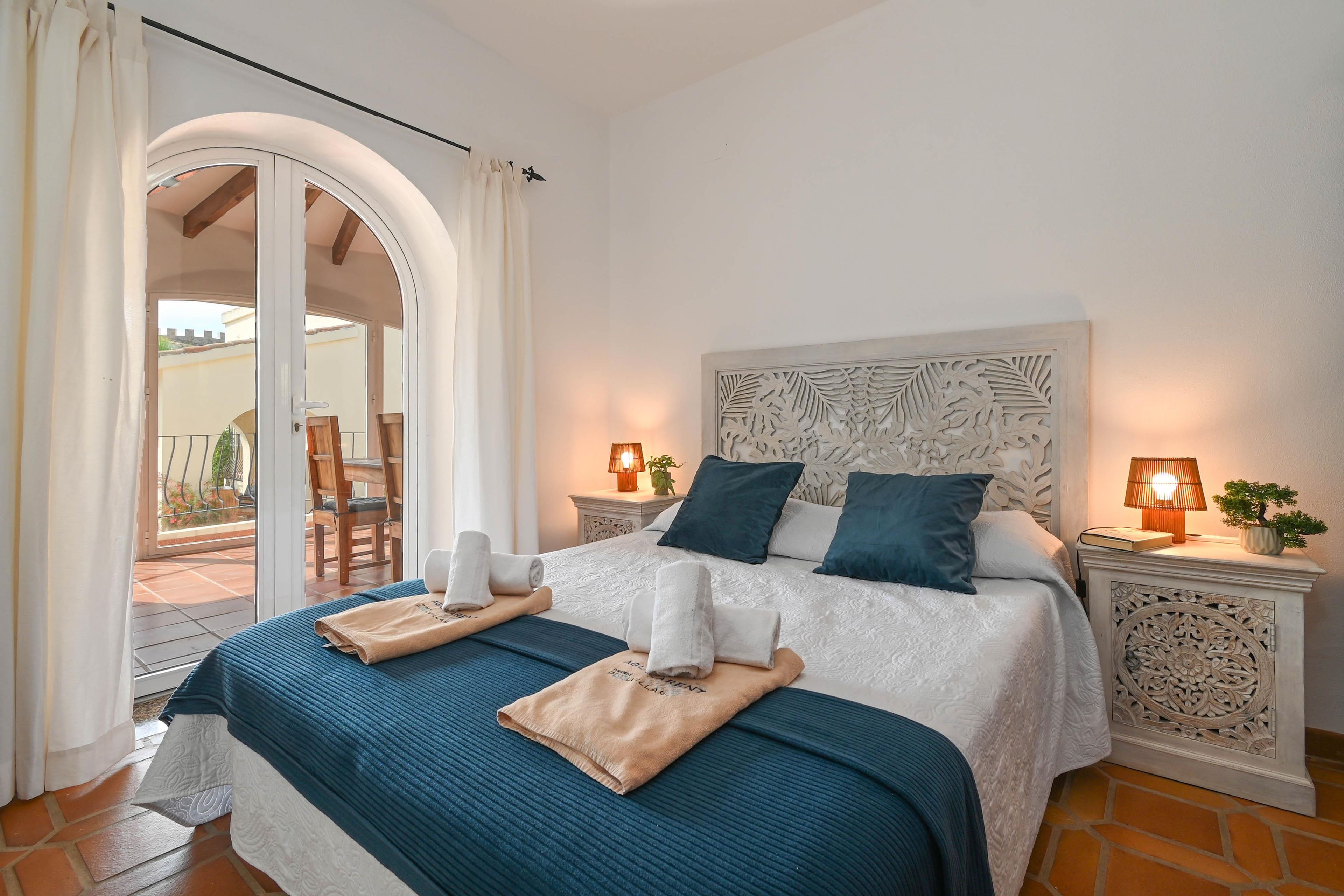A hotel room with a bed, two bedside lamps, a potted plant, and a view of the outside balcony.