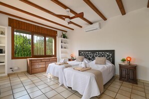 A bedroom with two beds, a wooden ceiling, a window with a view of greenery, and a wall-mounted air conditioner.