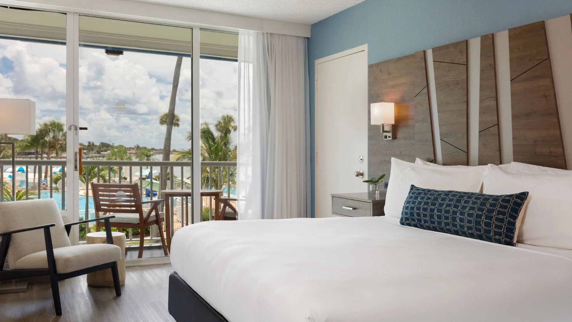 A hotel room with a large bed, a chair, a small table, a nightstand, and a view of a pool and palm trees.