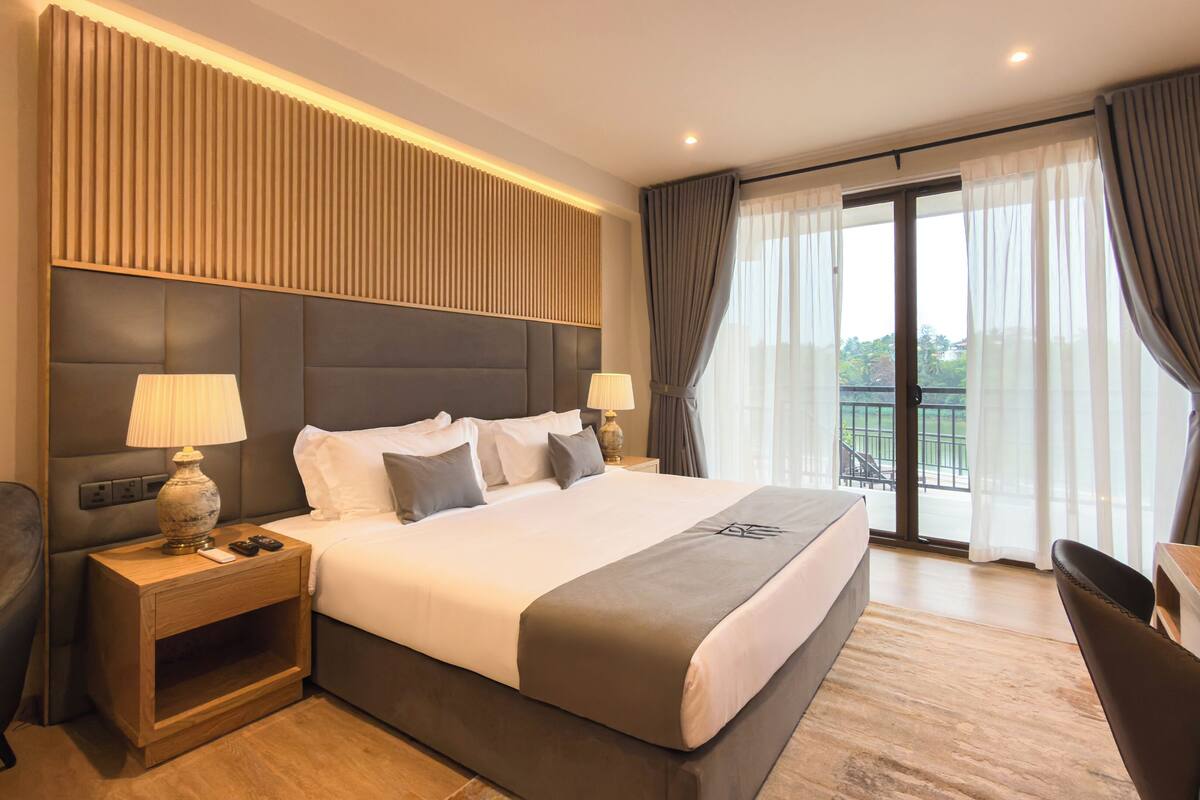 Habitación de hotel con una cama grande, dos mesitas de noche con lámparas, una silla y un balcón con cortinas.