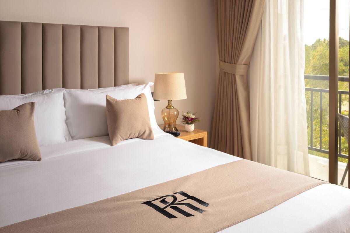 Una cama bien hecha con colcha y almohadas color beige, una lámpara de noche y vistas al verdor por la ventana.