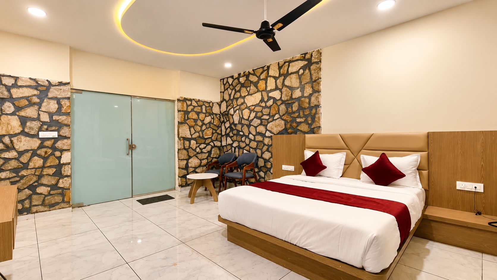 A hotel room with a stone accent wall, a bed with white bedding and red pillows, a ceiling fan, and a small table with chairs.