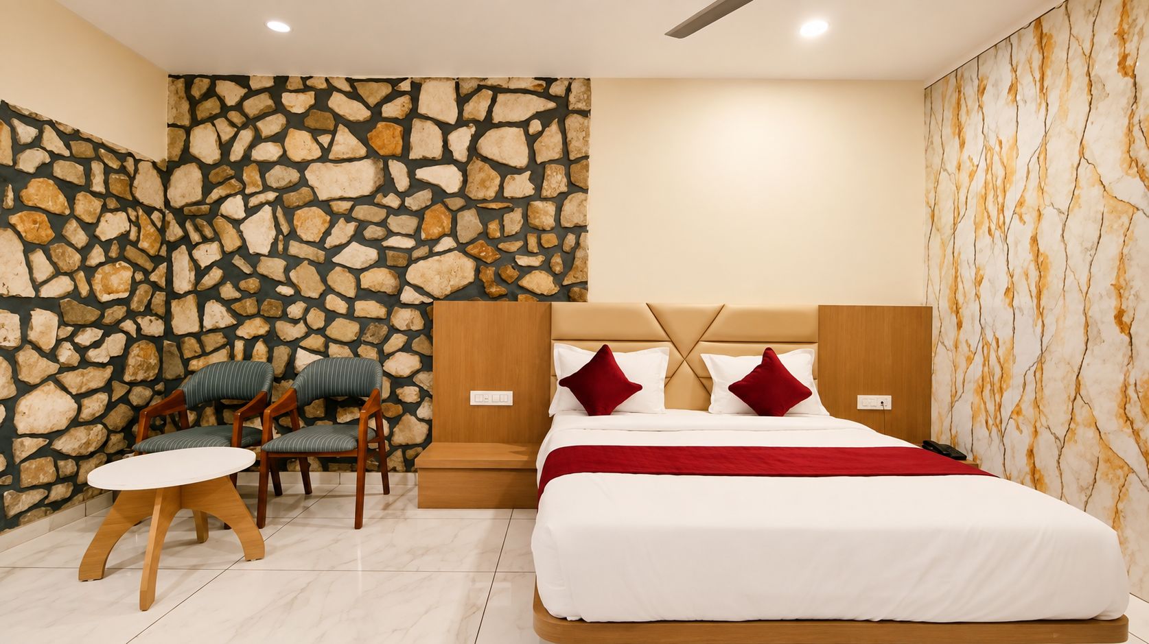 A hotel room with a stone wall, a bed with red pillows, a wooden nightstand, and a small table with chairs.