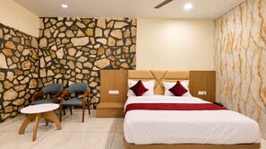A hotel room with a stone wall, a bed with white bedding and red pillows, a wooden headboard, a small wooden table, and two upholstered chairs. - Divydrashti Resorts (Waghoriya)