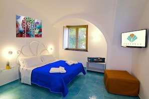 A hotel room with a bed, a window, a painting, a television, and a suitcase.