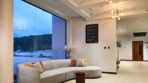 A modern hotel lobby with a curved gray sofa, a small round table, and a large window offering a view of a snowy landscape. - GOLDEN CASTLE (Neyyattinkara)