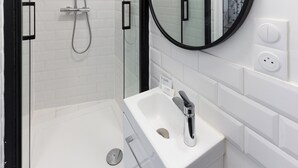 A modern bathroom with a glass-enclosed shower, a white sink, and a round mirror. - Studio 30s from RER E - Paris in 20 min! (Villemomble)