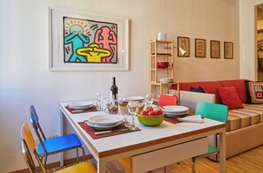 A dining area with a white table set for four, surrounded by colorful chairs. A framed abstract painting hangs above the table, and a wooden shelf with decorative items is visible in the background. - Charming and quiet two-room apartment in Porta Romana (Milano)