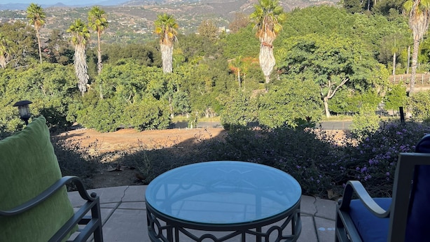 Une terrasse dotée d’une table en verre et de chaises en métal, offrant une vue sur un paysage arboré et des montagnes lointaines.