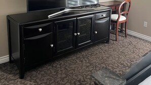 Une chambre d’hôtel dotée d’un grand téléviseur à écran plat fixé au mur, d’une commode en bois noire avec des tiroirs, d’une chaise en bois et d’un tapis à motifs.