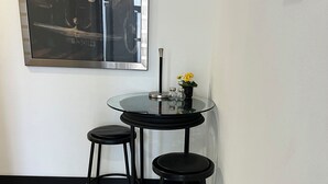 Une pièce d’angle avec une table en verre et deux tabourets, une œuvre d’art encadrée au mur et une petite plante en pot.