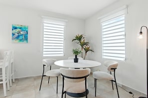 A dining area with a round table, six chairs, two potted plants, and a painting on the wall.