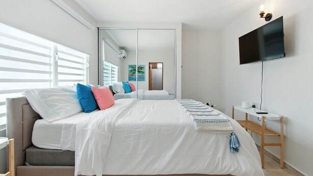 A modern bedroom with a large bed, a TV mounted on the wall, and a small table with a plant. - New 3-BR Apartment, A/C, Free Parking, Washer Dryer, Full Kitchen, WiFi (San Juan)