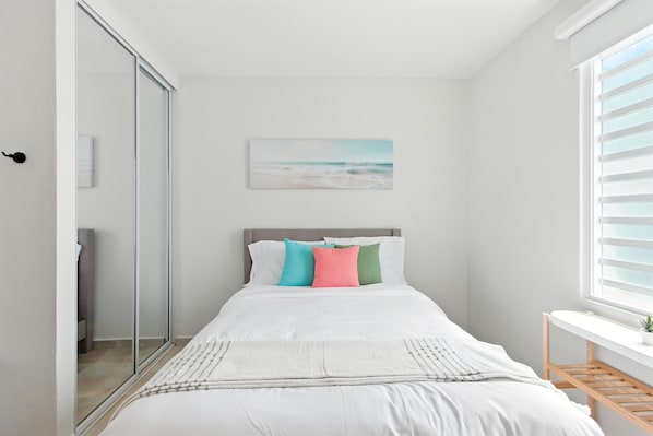 A bedroom with a bed, a window with blinds, a bedside table, and a painting on the wall.