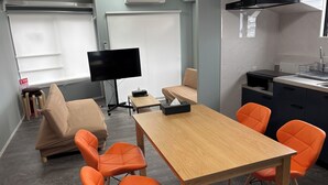 Una habitación moderna con una mesa de madera, sillas color naranja, un sofá, un televisor de pantalla plana y una zona de cocina con fregadero y estufa.