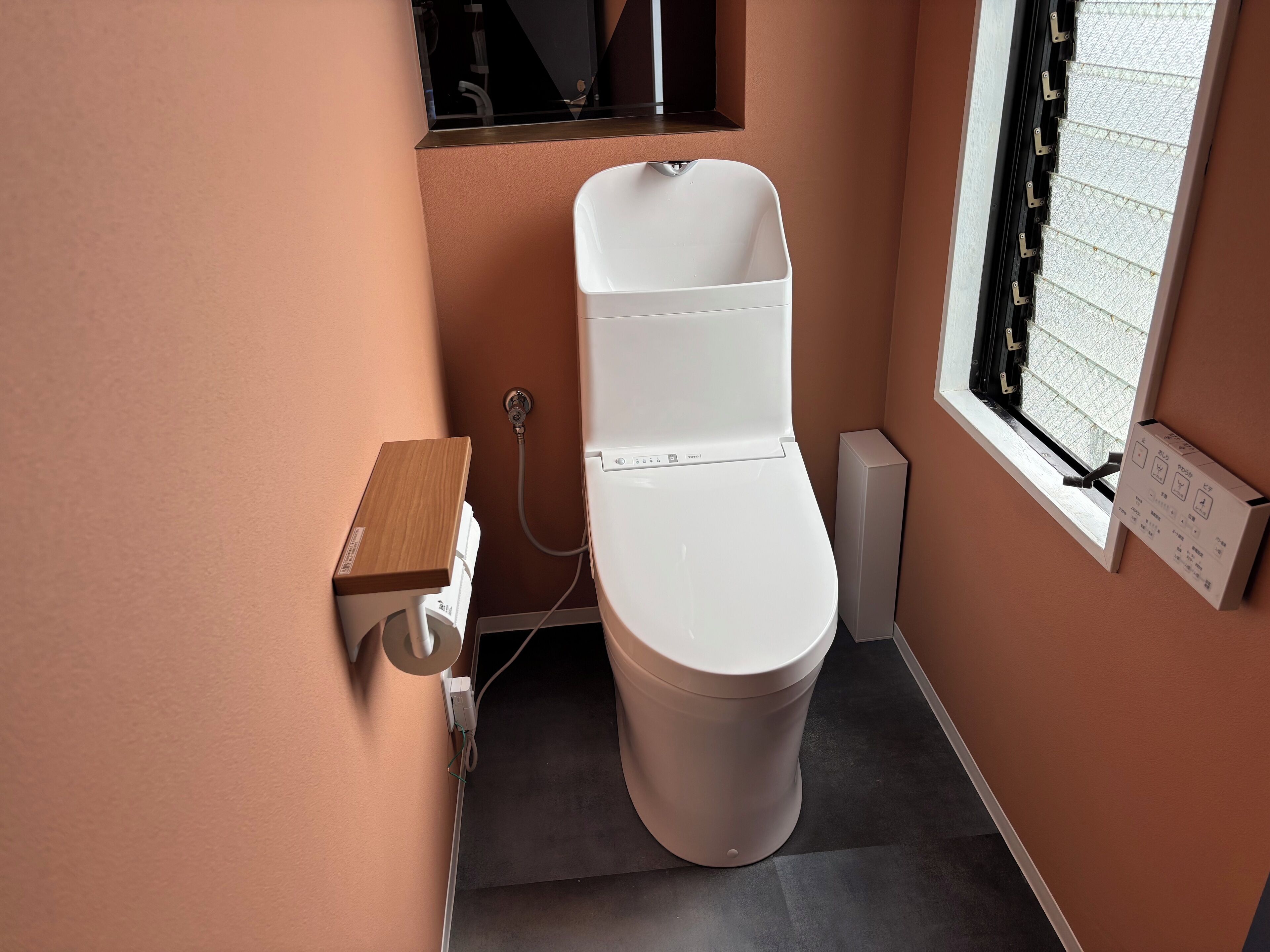 Une salle de bain moderne avec des toilettes blanches, une petite étagère en bois et une fenêtre avec une grille métallique.
