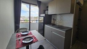 A compact kitchen with a dining table set for two, a large window with curtains, and a view of buildings outside. - Blue Edge Studio T3-527 (Chalong)