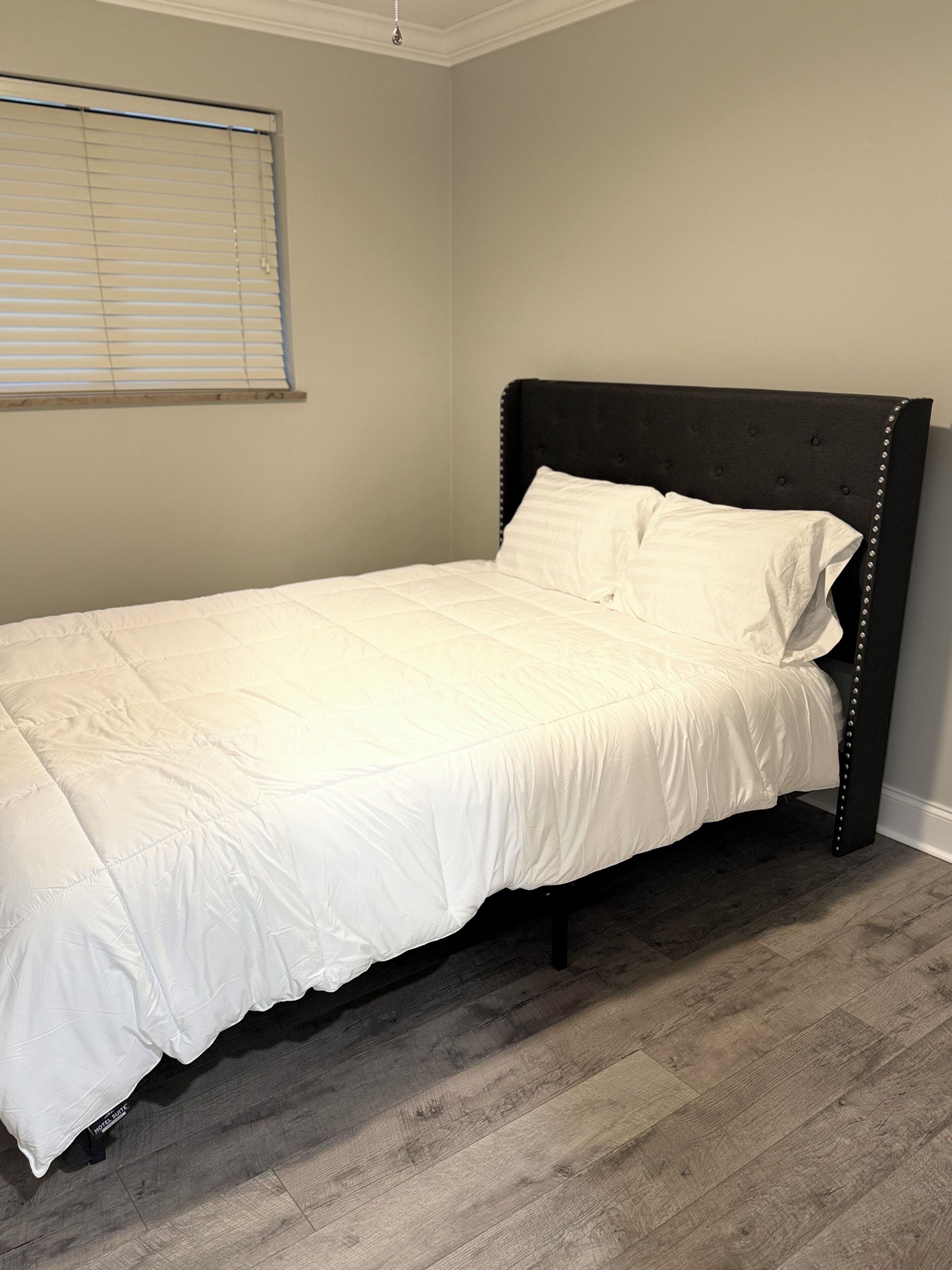 A single bed with a dark headboard and white bedding in a room with wooden flooring and a window with blinds.