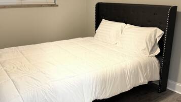 A single bed with a dark headboard and white bedding in a room with wooden flooring and a window with blinds.
