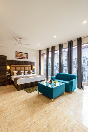 A modern hotel room with a large bed, a blue sofa, a glass coffee table, and a city view through large windows. - Hotel The Kiara - Complimentary Oneway Airport Transfer (New Delhi)
