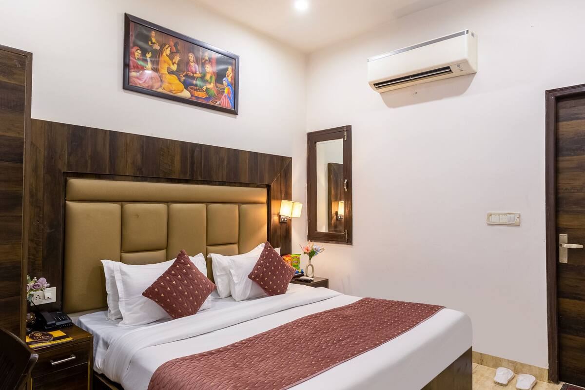 A hotel room with a large bed, wooden headboard, white bedding, a ceiling fan, an air conditioning unit, a framed painting, and a nightstand with a lamp.