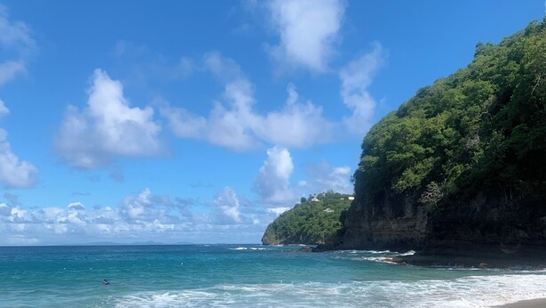 Une plage à l’eau d’un bleu limpide, avec une rive sablonneuse et une falaise couverte de végétation.