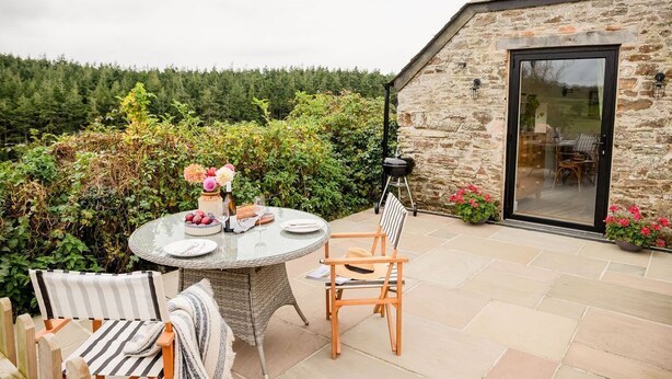 Een stenen gebouw met een glazen deur, een ronde tafel met stoelen, en een tuin met planten en bloemen.