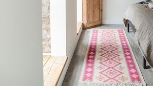 Een slaapkamer met een houten bed, een roze vloerkleed met patroon, een houten kastje met bloemen en een klok aan de muur.