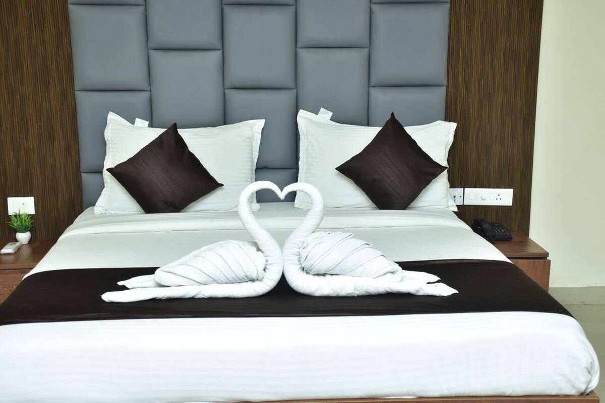 A hotel room with a tufted headboard, a bed with white and dark pillows, and two white swan-shaped towels on the bed.