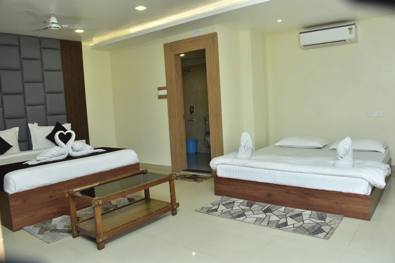 A hotel room with two beds, a wooden bedside table, a ceiling fan, an air conditioning unit, and a shower area.