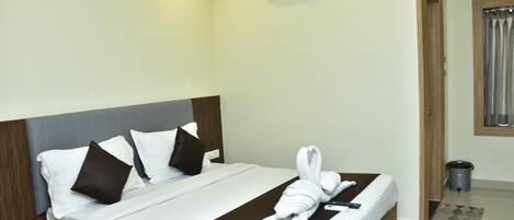 A hotel room with a bed, pillows, a towel, a remote control, an air conditioning unit, a door, and a trash bin.