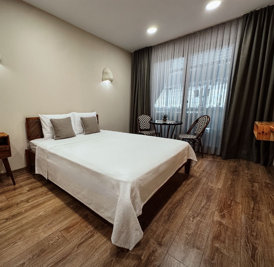 Habitación de hotel con una cama grande, mesitas de noche, una pequeña mesa redonda con sillas y una ventana con cortinas.