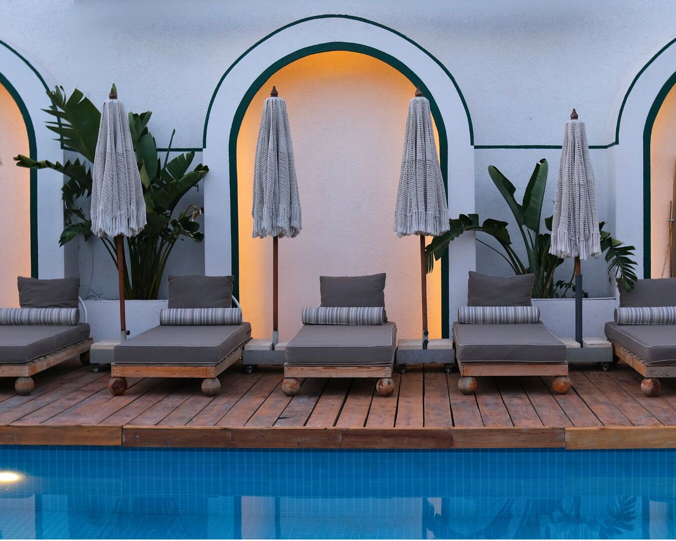 A poolside area with lounge chairs and umbrellas, surrounded by large plants and a pool.