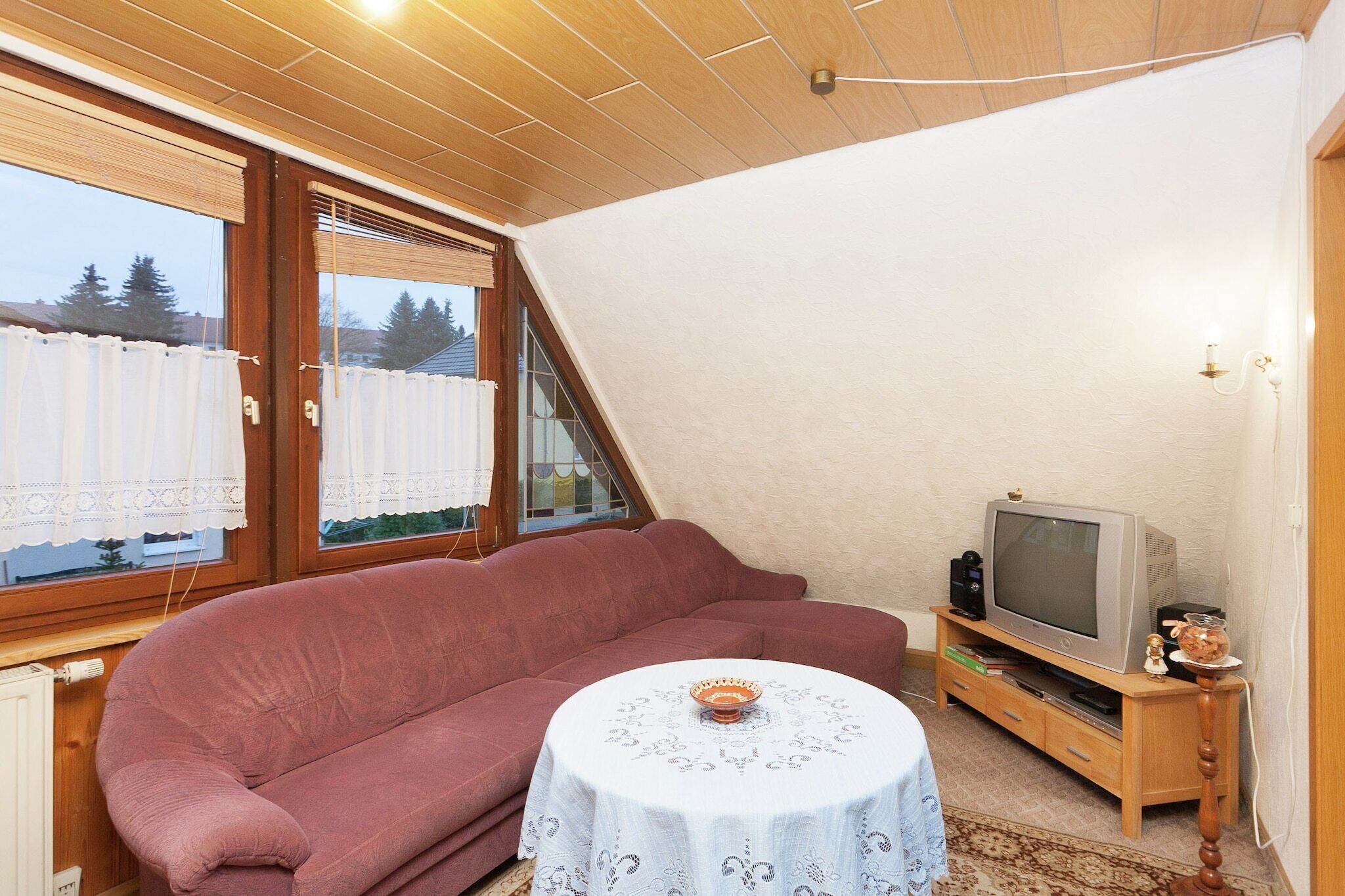Ein gemütliches Wohnzimmer mit einem roten Sofa, einem kleinen runden Tisch mit weißer Tischdecke, einem Fernseher auf einem Holzschrank und großen Fenstern mit Jalousien.