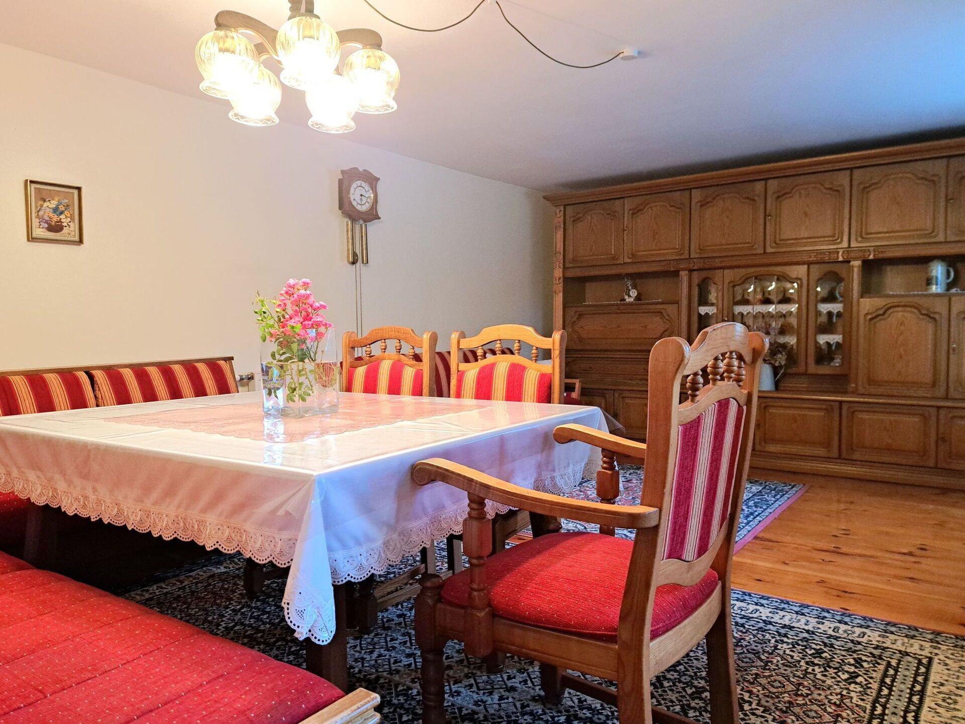 A dining area with a table set for four, surrounded by wooden chairs with red cushions. A vase with flowers is on the table, and a clock hangs on the wall.
